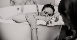 A woman giving birth in the bathtub while another woman is touching her arm, comforting her 