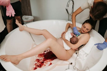 Young black-haired woman holding her newborn baby in the bathtub that is covered in blood