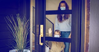 A young boy and his mother wearing masks and looking out the front door during the COVID-19 home qua...