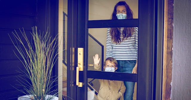 A young boy and his mother wearing masks and looking out the front door during the COVID-19 home qua...
