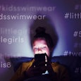 A young teenager is sitting in her bed in the dark with only the light of her phone shining on her f...