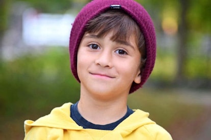 A young child in a yellow top and purple hat smirks.