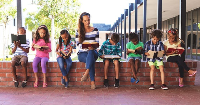Teacher and students reading book while sitting on brick wall at corridor