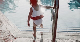 young girl at the side of an outdoor swimming pool, wearing frilly swimsuit, dipping toes into the w...