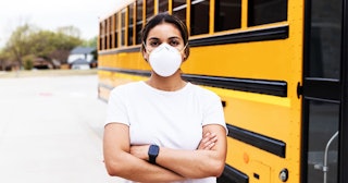 woman wearing an N95 mask, stands next to a school bus.
