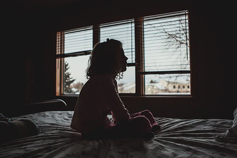 Little girl sits on a bed in a dark bedroom with blue sky and trees can be seen behind her in the wi...