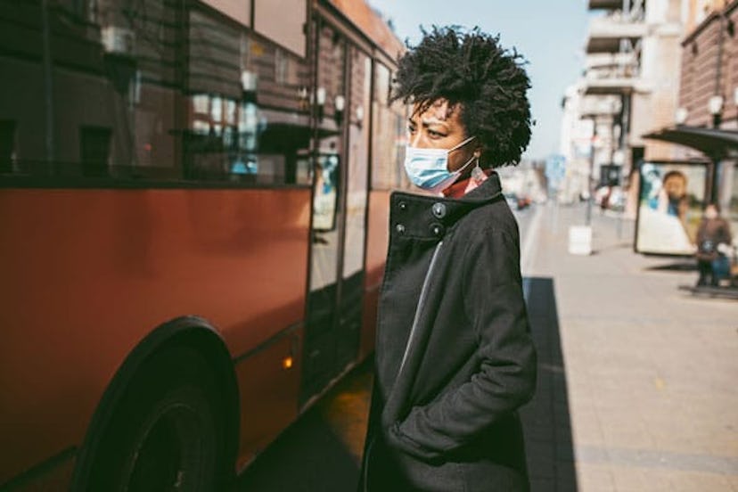 woman standing on city street with protective mask on her face and waiting for bus