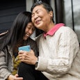 Elderly mother wearing a white sweater smiling while hugging her daughter on the porch of their hous...