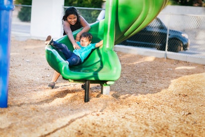 Older sister helping her younger brother go down a twisted slide in a kids playground