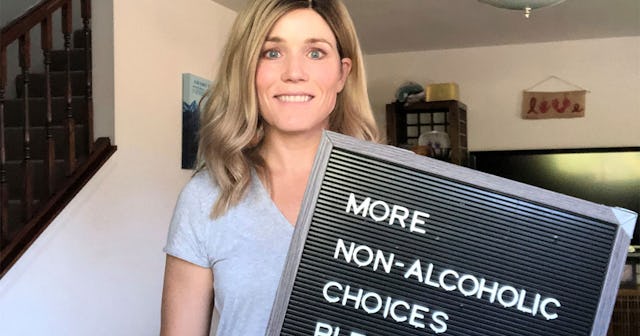 A blonde, blue-eyed woman in a light blue T-shirt holding a black sign with the words 'More Non-Alco...