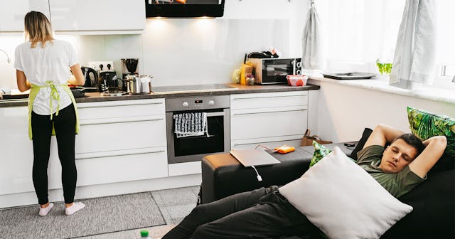 Woman Cooking In The Kitchen While her Lazy Husband Is Laying On The Couch