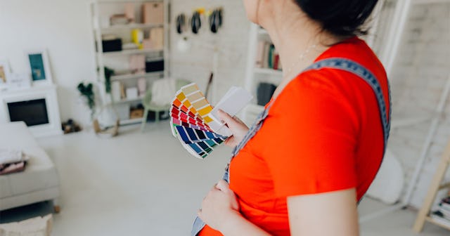 A pregnant woman in a red shirt holding a color shade swatch book for her baby's nursery redecoratio...