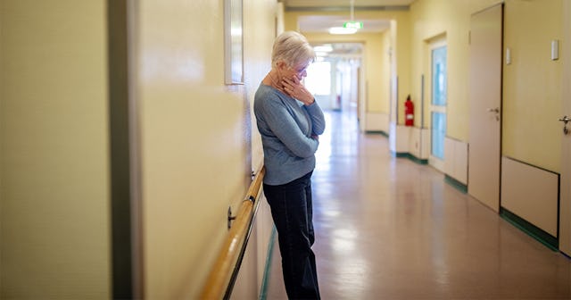 An older woman with gray hair, wearing blue clothes, standing in a nursing home hallway