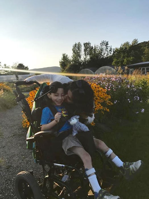 A happy mother kissing her smiling eight-year-old son, sitting in his wheelchair outside on a sunny ...