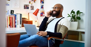 A man with a 'work laugh' sitting with his laptop on his lap and speaking on a phone while smiling