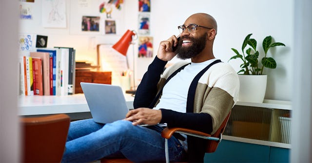 A man with a 'work laugh' sitting with his laptop on his lap and speaking on a phone while smiling