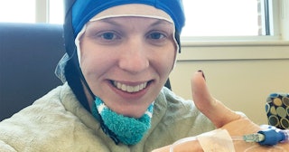 Megan Gill Carusona who is battling breast cancer, smiling in a hospital and holding one thumb up