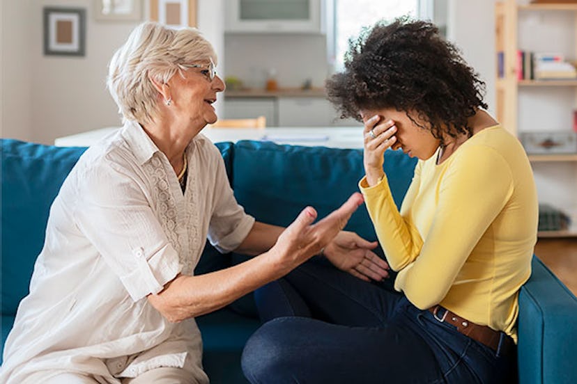 A mom comforting her sad grown-up daughter while they're sitting on a white couch in a living room