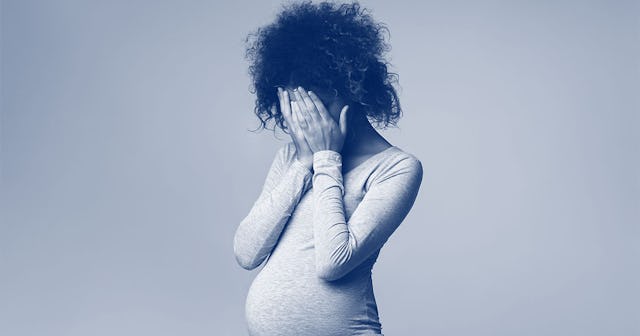 Pregnant woman covering her face while standing in front of a wall.