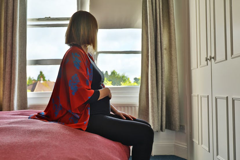 A pregnant woman sitting on a bed looking out of the window.
