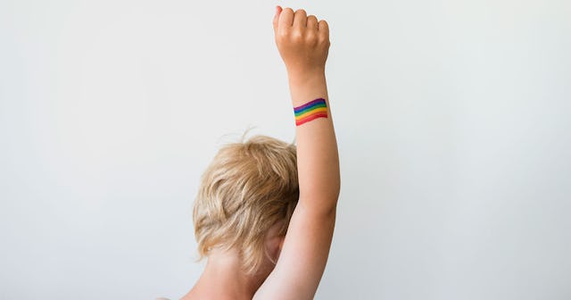 A transgender child standing with his right arm in the air and a rainbow flag painted on his wrist.
