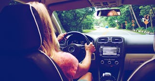 The back of a woman driving her car through a street with green trees on the side of the street