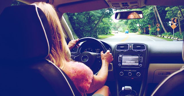 The back of a woman driving her car through a street with green trees on the side of the street