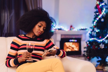 A curly-haired woman sitting alone in a festive living room with a Christmas tree and drinking wine ...