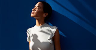 A dark-haired woman in a white top with frilled sleeves with closed eyes in front of a navy-blue wal...