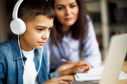 A boy typing on his laptop with headphones on, while his mom is sitting next to him during homeschoo...