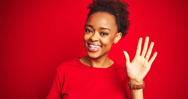 A woman with an Afro hairstyle wearing a red dress in front of a red wall, smiling and waving farewe...