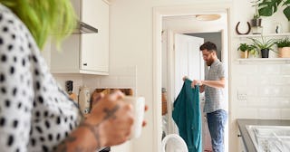 A wife in a white shirt with black dots drinking coffee while watching her husband is taking the lau...