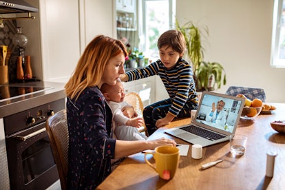 Red-haired mother with her toddler and her older child sitting on a table with a laptop on it, talki...