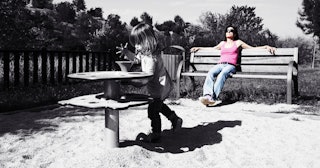 A woman sitting on a bench at a playground while a girl is playing in front of her.