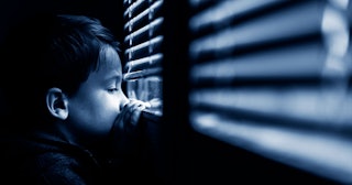 Depressed-looking boy leaning against the window and looking bellow the shutters outside from a dark...