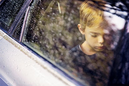 Depressed-looking blonde boy staring at the floor of a car while driving in it with the rain falling...
