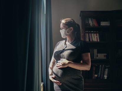 Pregnant woman with face mask standing in front of window.