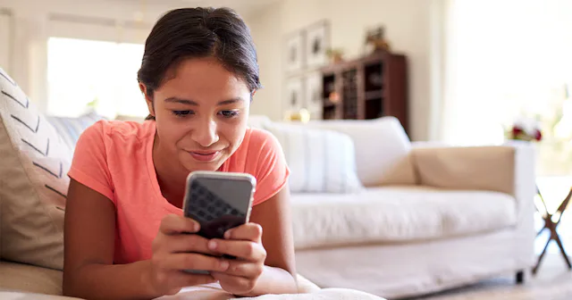 An 8th grade girl with a smart phone in her hands while lying stomach down a beige