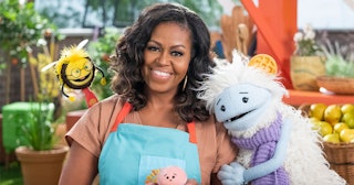 Michelle Obama, wearing a blue apron, stands in a rooftop garden posing for the camera and holding a...