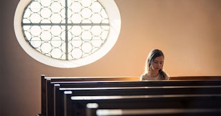 A girl sitting in the last row pew in a church with her head down and a large mirror behind her