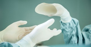 A person's hand in white gloves giving a breast implant to another doctor during a surgery 
