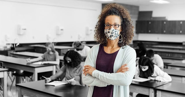 A teacher wearing a mask in the classroom, her students taking a test behind her.