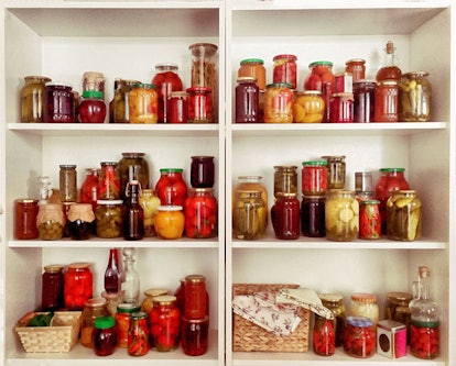 Six sections of an open kitchen cabinet filled with jars containing various kinds of pickled vegetab...