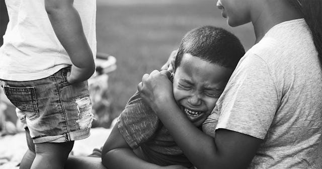 A child crying in his mother's arms and another child standing next to them in black and white