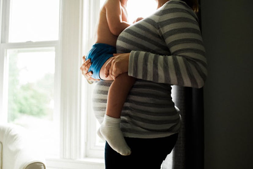 A pregnant woman in a grey-and-white striped shirt holding a baby in blue shorts and white socks abo...