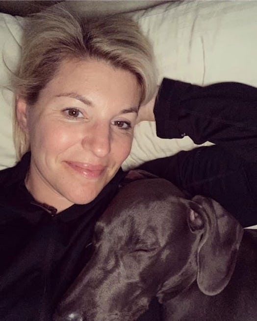 A woman lying in a bed with one hand under her head with a dog sleeping on her chest.