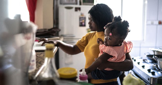 A black SAHM wearing a yellow t-shirt holding her daughter in arms in the kitchen during a pandemic