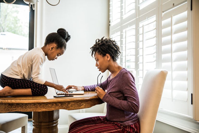 A black SAHM working on her laptop while her daughter is sitting and using her smartphone during the...