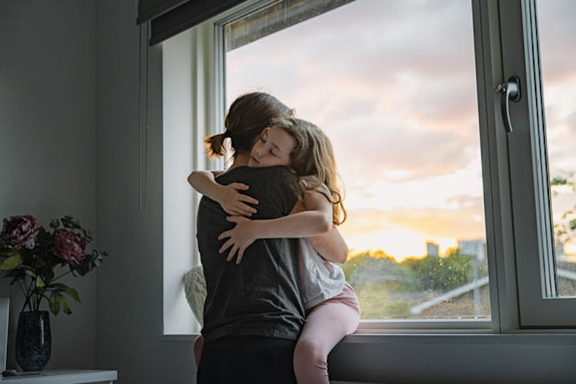 A mother hugging her daughter sitting at a window
