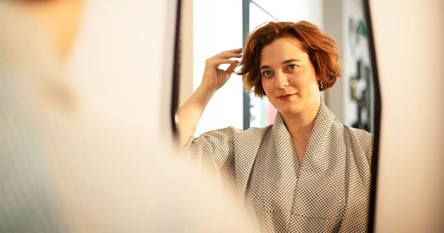 A woman with short brown hair looking into an oval mirror on the wall